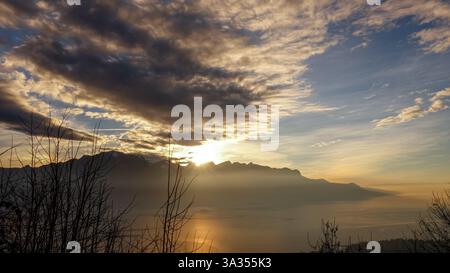 Atemberaubender Blick auf den Sonnenuntergang über dem Genfer See mit der Silhouette der französischen Alpen im Hintergrund, unter einem dynamischen Wolkenhimmel. Stockfoto