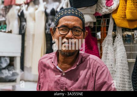 Ein balinesischer Mann in einem rosafarbenen Hemd lächelt, während er an einem Marktstand vor bunten Häkeltaschen steht und verschiedene handgefertigte Handwerkskunst präsentiert. Stockfoto