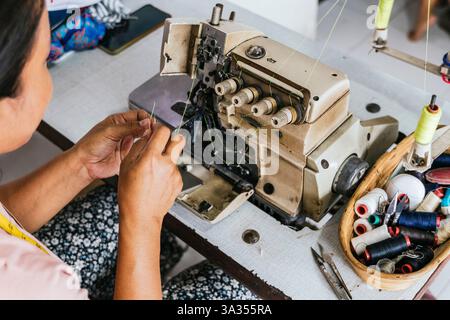 Eine Nahaufnahme einer asiatischen Schneiderin, die an einer Nähmaschine arbeitet, umgeben von bunten Fadenspulen, die die Kunst des Schneidens widerspiegeln Stockfoto