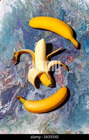 Drei hellgelbe Bananen, eine davon geschält, ruhen auf einem lebendigen abstrakten Marmorhintergrund. Die Farben sorgen für einen auffälligen Kontrast, der die Frucht hervorhebt Stockfoto