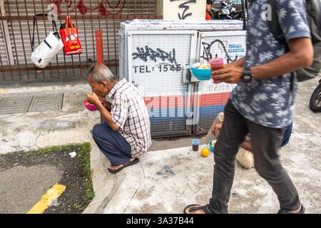 Ein älterer Mann, der sich hockt und einen Essensbecher hält. Eine Person, die mit einer Tasse Essen läuft, Straßenszene, Chinatown Kuala Lumpur, Malaysia, Asien Stockfoto