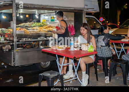 Eine Nachtmarktszene, Kuala Lumpur Malaysia, mit einem Lebensmittelhändler, der verschiedene Gerichte zubereitet und serviert. Allein eine Frau, die am Tisch isst Stockfoto