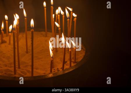 Die Kerzen sind in Sand angeordnet und ihre Flammen leuchten warm vor einem dunklen kirchlichen Hintergrund, was Gefühle von Spiritualität, Gelassenheit und Reflexion hervorruft Stockfoto
