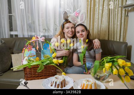 Mutter und Tochter mit Hasenohren lächeln, während sie gelbe Tulpen in gemütlicher Osterstimmung halten. Dekorierter Korb, handgefertigtes Kunsthandwerk und glasierte Kuchen Stockfoto
