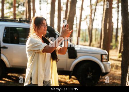 Die schöne Frau steht im Wald mit dem Smartphone in den Händen. Gegen modernes Auto. Stockfoto