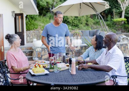 Eine Gruppe verschiedener älterer Freunde genießt ein Mittagessen im Freien, lächelt und plaudert um den Tisch herum Stockfoto