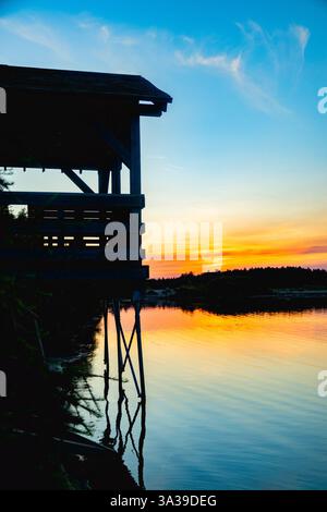 Ein wunderschöner Sonnenuntergang mit der Silhouette einer Hütte am See, die eine friedliche und ruhige Atmosphäre schafft. Stockfoto