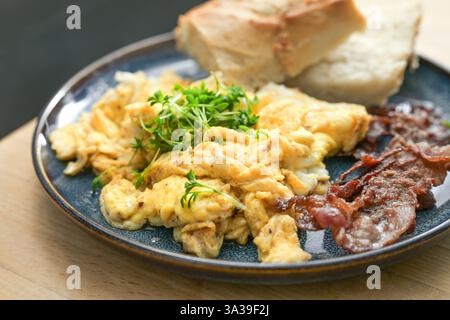 Rührei mit Kresse-Beilage, knusprig gerösteter Speck und Brot und auf einem Teller auf einem Holztisch, herzhaftes Frühstück, Mittag- oder Abendessen, ausgewählte Fokussierung, Stockfoto