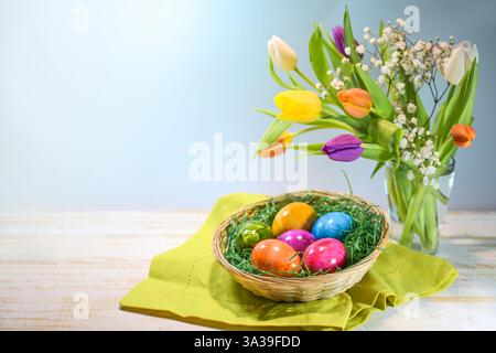 Bunte Ostereier in einem Korb und ein Frühlingsstrauß mit Tulpen auf hellem Holz vor blauem Hintergrund, Weihnachtsessen und Dekoration, Kopierraum, se Stockfoto