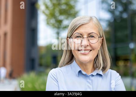 Ein Porträt einer Reifen, selbstbewussten Geschäftsfrau, die draußen lächelt, mit moderner Architektur und Grün im Hintergrund. Stockfoto