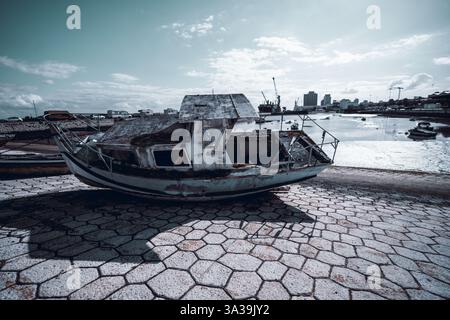 Ein verrostetes, verlassenes Fischerboot mit einem verrosteten, gebrochenen Rumpf und zersplitterten Fenstern sitzt an einem gepflasterten Ufer in der Nähe eines Hafens. Städtische Skyline, Kräne und Boa Stockfoto