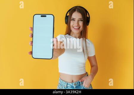 Kopfhörer auf dem Kopf, für das Smartphone mit Kopierraum. Die junge Frau ist auf gelbem Hintergrund. Stockfoto