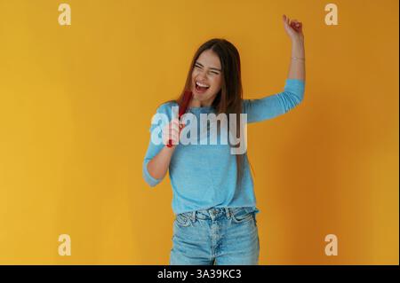 Singen in die Haarbürste. Die junge Frau ist auf gelbem Hintergrund. Stockfoto