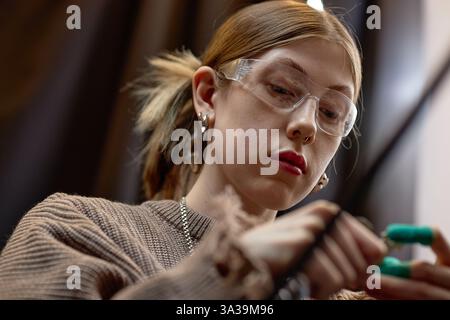Nahaufnahme einer fokussierten Frau, die eine Schutzbrille trägt und in einer Werkstattumgebung arbeitet und sich mit Präzision und Aufmerksamkeit auf eine Aufgabe konzentriert Stockfoto