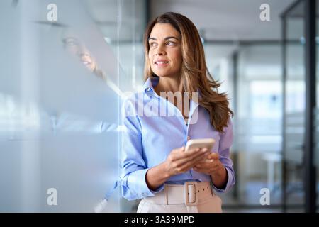 Reife Geschäftsinvestorin, die ihr Telefon benutzt, im Büro steht und wegschaut. Stockfoto