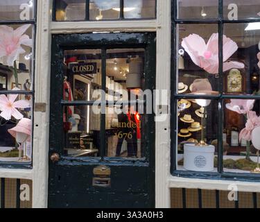 Frontfenster eines hübschen Hutgeschäfts aus dem 17. Jahrhundert, während sich zwei Männer im Inneren unterhalten und einer eine Flamme angezündet hat. London, März 2025 Stockfoto