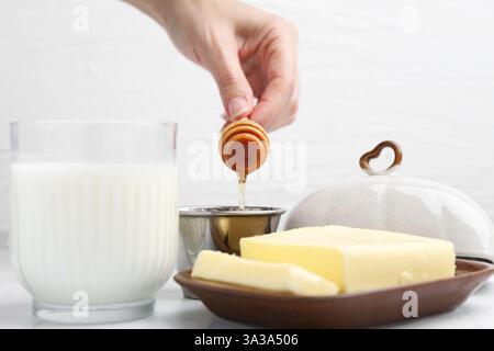 Frau mit Honiglöffel, Nahaufnahme. Frische Milch im Glas und Butter auf weißem Fliesentisch Stockfoto