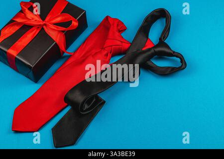 Rote und schwarze Krawatten liegen auf einer blauen Oberfläche neben einer wunderschön verpackten Geschenkbox. Stockfoto