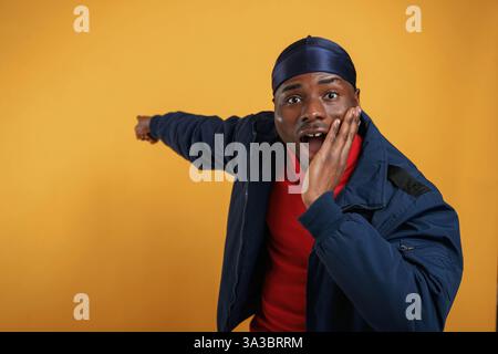 Schockiert, mit der Hand etwas an der Wand zu zeigen. Ein hübscher schwarzer Mann ist im Studio vor gelbem Hintergrund. Stockfoto