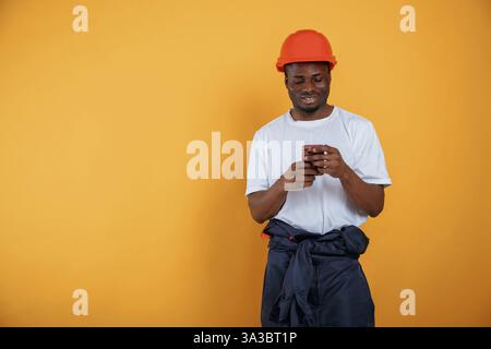 Fröhlich, Smartphone in der Hand, mit Schutzhelm. Ein hübscher schwarzer Mann ist im Studio vor gelbem Hintergrund. Stockfoto