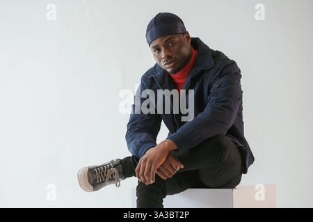 Sitzt auf dem Würfelstuhl. Schwarzer Mann mit Kappe ist im Studio vor weißem Hintergrund. Stockfoto