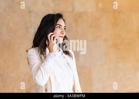 Eine professionelle Frau in eleganter Geschäftskleidung spricht mit einem durchdachten Ausdruck auf ihrem Smartphone. Sie steht vor einer modernen Steinmauer, verlobt Stockfoto