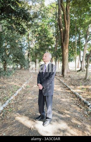 Ein Park an einem angenehmen Frühlingstag mit viel Grün in Japan. Ein Japaner in den späten 70ern, der einen Anzug trägt, steht auf. Stockfoto