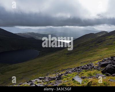 Dunkle Wolken überragen eine bergige Landschaft. Sonnenlicht bricht durch und beleuchtet Täler und zwei Seen. Stockfoto