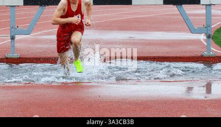 Ein Athlet in hellen Schuhen navigiert bei einem anspruchsvollen Rennen bei nassem Wetter über eine wassergefüllte Hürde. Stockfoto