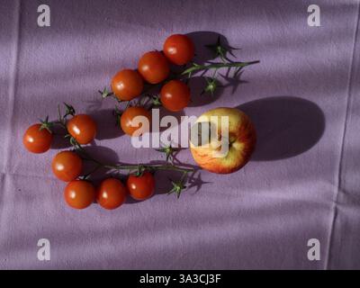 Kirschtomaten und ein Apfel auf violetter Tischdecke, Horizontalansicht von oben nach unten bei Sonnenlicht Stockfoto