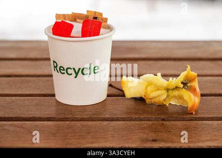 Ein mit „Recycle“ gekennzeichneter Papierbecher steht auf einem Holztisch, gefüllt mit Zuckerverpackungen, künstlichem Süßungsmittel und Apfelkern zum Recycling Stockfoto