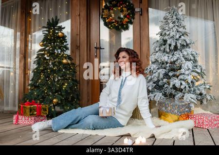 Frau Weihnachtsbäume dekorierte Veranda lächelnde Frau auf einer Veranda sitzend dekoriert für Weihnachten mit zwei Bäumen und Geschenken. Stockfoto