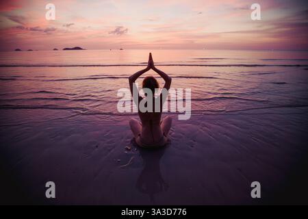 Eine Frau sitzt in Lotusposition, meditiert während eines ruhigen Sonnenuntergangs am Ufer des Wassers und verbindet Ruhe und natürliche Schönheit. Stockfoto