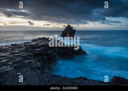 Wellen krachen gegen zerklüftete Felsformationen, während die Dämmerung ein atemberaubendes Licht über die Szene zaubert. Der Himmel ist voller Wolken, die das natürliche B verstärken Stockfoto