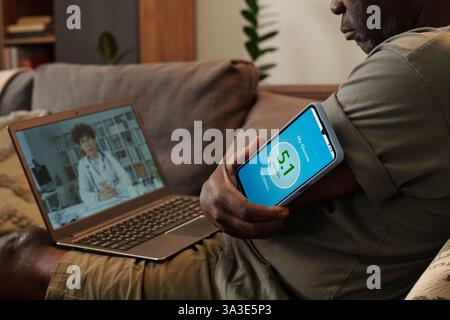 Älterer Mann mit Online-Arzttermin mit Arzt sichtbar auf Laptop-Bildschirm während Verwaltung der Gesundheits-App auf Mobiltelefon Stockfoto