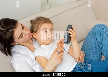 Mutter und Tochter teilen einen verspielten Moment zusammen auf der Couch. Die Mutter, die eine weiße Bluse und Perlenzubehör trägt, berührt sie sanft Stockfoto