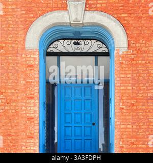 Leuchtend blaue Holztür mit kompliziertem eisernem Heckspiegel, eingerahmt von einem historischen roten Backsteinbogen und Steindetails. Stockfoto