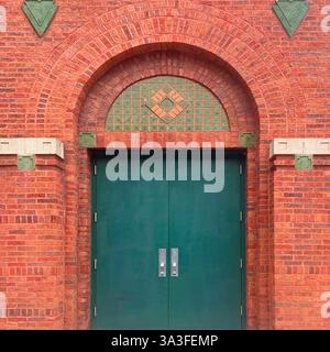 Grüne Doppeltüren in einer bogenförmigen roten Ziegelfassade mit geometrischen Ziegelmustern und dekorativen grünen Flieseneinlagen. Stockfoto