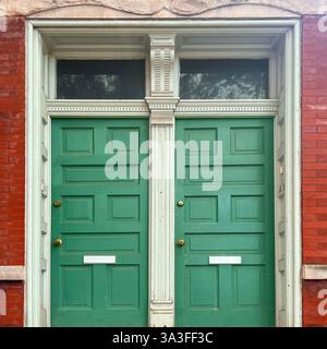 Alte grüne Doppeltüren mit weißen dekorativen Zierleisten und Heckfenstern, die vor einer historischen roten Backsteinfassade stehen. Stockfoto