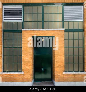 Eingang zum Industriebau mit grüner Metalltür, eingebettet in eine gelbe Ziegelfassade, eingerahmt von großen Gitterfenstern mit Lüftungsblechen. Stockfoto
