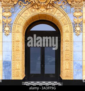 Ein reich verzierter bogenförmiger Eingang mit Bogentüren, umgeben von aufwändigen goldgelben Terrakotta-Details, blauen Fliesen und kariertem Boden. Stockfoto