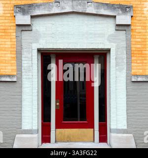 Eine rote Tür mit dekorativen Säulen und einer Fußplatte aus Messing, eingerahmt von einer gedämpften Ziegelfassade in Gelb- und Grautönen, die eine Vintage-Architektur darstellt Stockfoto