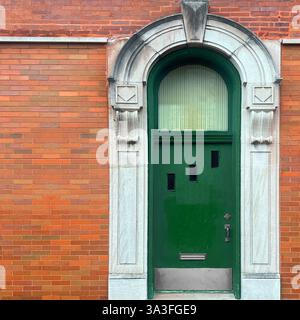 Tür mit einer bogenförmigen grünen Holztür mit einzigartigen asymmetrischen Fenstern, eingerahmt von einer lebendigen orangen Ziegelfassade mit Steinschnitzereien. Stockfoto