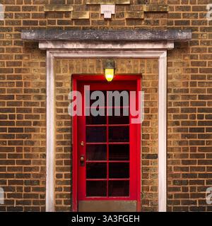 Historische Stadttür mit roter Tür mit Glasscheiben, beleuchtet von einer warmen gelben Laterne, eingerahmt von detailliertem Mauerwerk und Ziegelfassade. Stockfoto