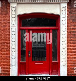 Historische Stadttür mit einer lebendigen roten Holztür mit dekorativen Glaspaneelen, elegant eingerahmt von detaillierten Steinverkleidungen und Ziegelfassade. Stockfoto