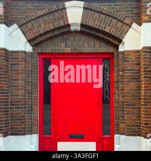 Rote Tür vor einer braunen Ziegelfassade, eingerahmt von einem dramatischen gotischen Bogen mit kontrastierenden Steinakzenten. Stockfoto