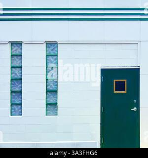 Eine minimalistische grüne Tür mit einem kleinen Fenster vor einer weißen geometrischen Fassade mit vertikalen Glasblockfenstern und Art déco-Linien. Stockfoto