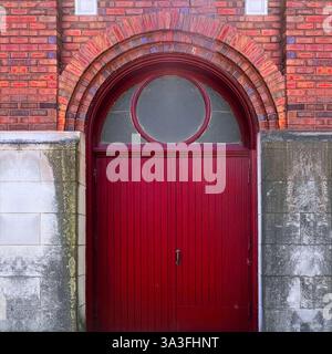 Eine historische rote Tür mit einem bogenförmigen Heckfenster, eingebettet in eine Ziegelstein- und Steinfassade, Eingang mit halbrunder Glasscheibe Stockfoto