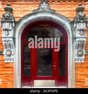 Elegante rote Tür, eingerahmt von einem komplizierten Steinbogen, der mit Löwenköpfen und dekorativen Elementen geschmückt ist, vor einer warmen Ziegelfassade Stockfoto