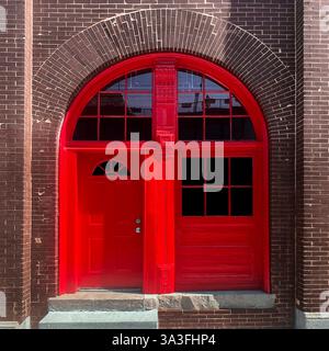 Hellrote Bogentür mit angrenzenden Fensterpaneelen in einem braunen Backsteingebäude mit einem markanten Kontrast zwischen Architektur und Farbe Stockfoto